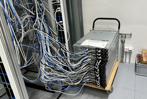 Auf einem Kurier-Wägelchen stehen rund 14 eingeschaltete, mit mehreren Ethernet-Kabeln verdrahtete Server neben einem Rack.
