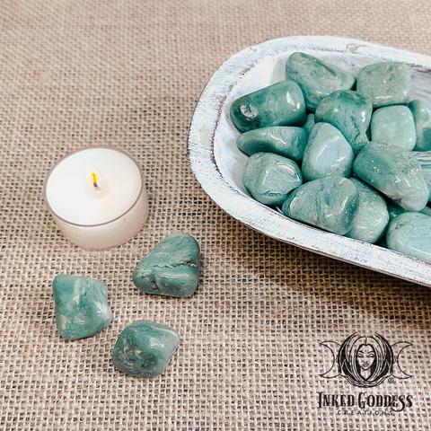 Three specimens of our "Mtorolite Tumbled Gemstone" product are arranged on the left in front of a tealight candle. Several more specimens fill a whitewashed wood bowl on the right. Presented by Inked Goddess Creations.