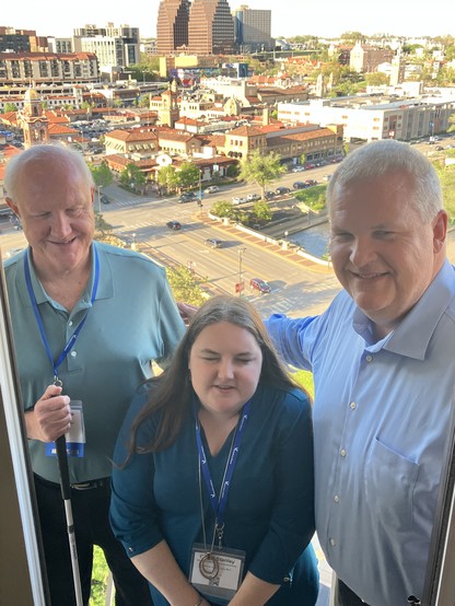 Photo of (left to right) ACB Special Projects Manager Dan Spoone, ACB Director of Advocacy and Governmental Affairs Claire Stanley, and ACB Executive Director Scott Thornhill standing on a balcony with the Kansas City skyline in the background. All three are smiling. It is late in the day and the sun is illuminating the buildings in a bright orange glow. 