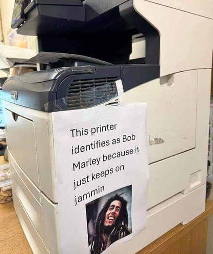 Image of an MFC Printer with a sign with an image of Bob Marley on it.
Sign text "This printer identifies as Bob Marley because it just keeps on jammin"