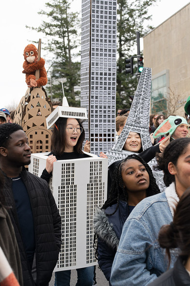 Numerous students in this portait-oriented images including two Asian girls inside building costumes as well as a stuff monkey on top of the empire state building.