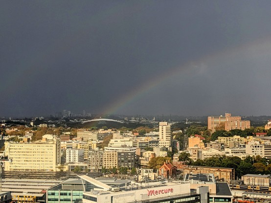 A semi-aerial photograph from the top floor of a tall building, looking out over Sydney's inner eastern suburbs. The sky is fully grey from a quick storm, and there's a large rainbow very close, apparently ending over some nearby apartment blocks. A faint double rainbow is visible. Landmarks in frame include Central Station in the foreground, the Sydney Football Stadium in the background, and Bondi Junction behind it.