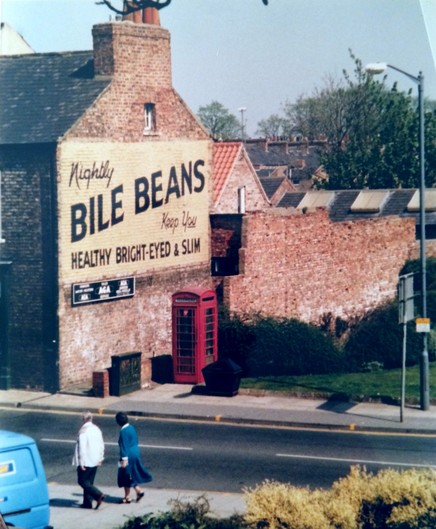 Slightly elevated view of an end-terrace house with a traditional red British phone box just in front of the gable-end wall which has a hand-painted sign across its entire width. The background of the sign is yellow and the lettering (quoted in the accompanying post) is in black.