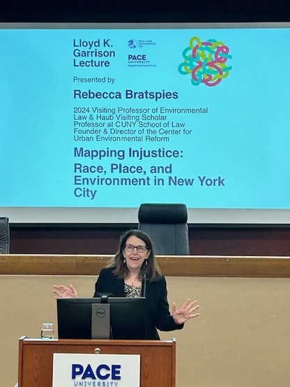 Me (a white woman with brown hair and glasses, wearing a black jacket) standing in front of a screen that announces the Lloyd K. Garrison Lecture. The title of my lecture is Mapping Injustice: Race, Place, and Environment in New York City.