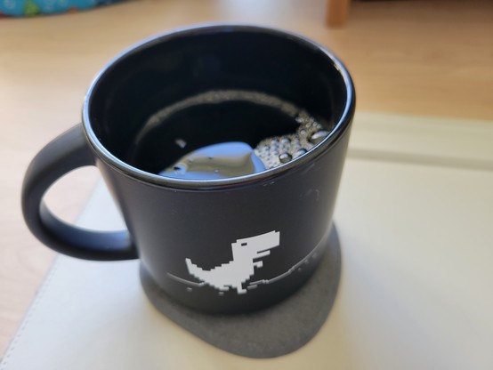 Black mug of cold brew coffee on a gray diatomaceous coaster on an off-white desk mat on a maple desk. The mug shows a pixelated dino in white on a black background. The dino is the protagonist T-Rex from The Dinosaur Game, a browser game developed by Google and built into the Google Chrome web browser.