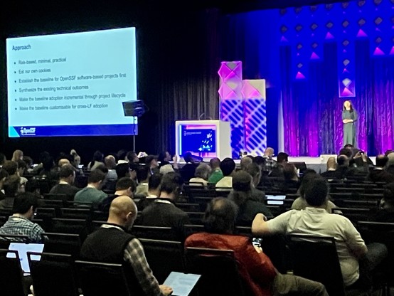 A conference presentation with an audience looking towards a speaker standing beside a large screen displaying bullet points.