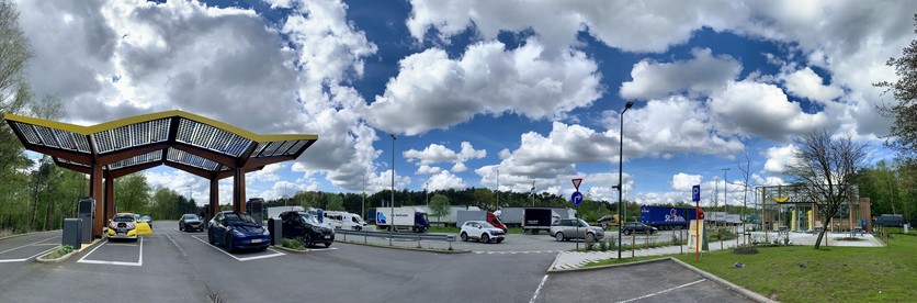 Panoramic view of a highway rest area with solar panel-covered carports, electric vehicle charging stations, parked cars, and trucks in the background, under a partly cloudy sky.