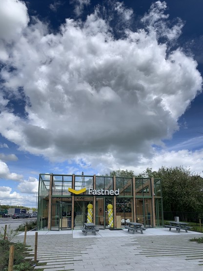 A modern glass building with the sign "Fastned" under a large sky with puffy clouds. There are yellow balloons inside and a yellow logo above the entrance. Benches are in front and a highway is visible in the background.