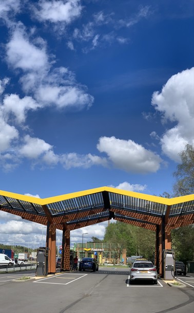 Solar panel canopy over electric vehicle charging stations with cars parked underneath, set against a blue sky scattered with fluffy clouds.