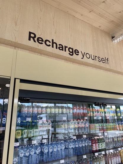 Refrigerated beverage display with a variety of drinks underneath a wooden panel with the slogan "Recharge yourself" printed on it.
