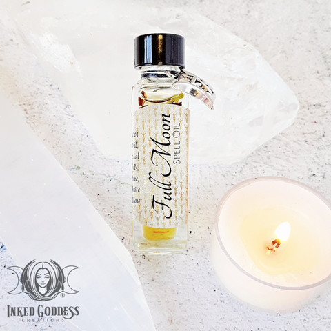 A small square vial of Full Moon Spell Oil. It has a silver triplemoon charm tied to the neck of the vial. It's laying against a chunk of white quartz with a selenite charging plate nearby and a white tealight candle for scale. Presented by Inked Goddess Creations.