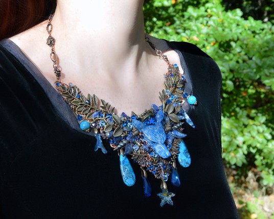 A large wire wrapped bib necklace, worn against a black velvet dress. The necklace is V shaped and features a bright blue crowned wolf in the centre made from Lapis Lazuli stone. Bronze leaves and blue crystals branch either side of him, with tiny seashells. Various pendants hang underneath - blue stone teardrops, Swarovski cystal starfish and blue beads.