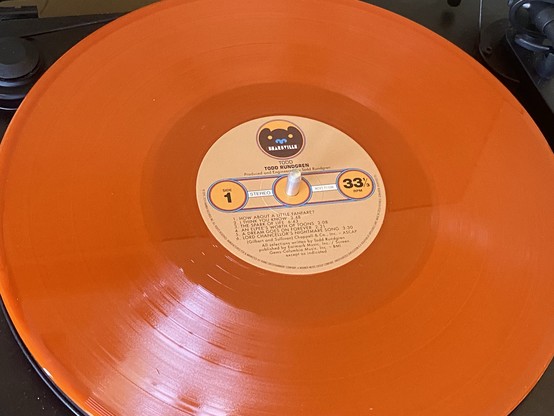 An orange vinyl record on a turntable. The center label shows "Todd Rundgren" and track listings for side 1.