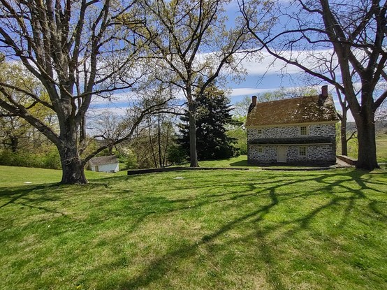 Photo: On the right, an old stone home with chimneys at either end and a brown roof, separated from some sort of white outbuilding with a single chimney that is farther in the background toward the left. A few dark green pine trees are at the middle of the scene. Several deciduous trees are just beginning to sprout leaves — three such trees are most prominent. The ground is covered with light green grass, and rolls gently away. The sky is bright blue and striped with clouds. Shadows from the trees gently dapple the sunlit grass.