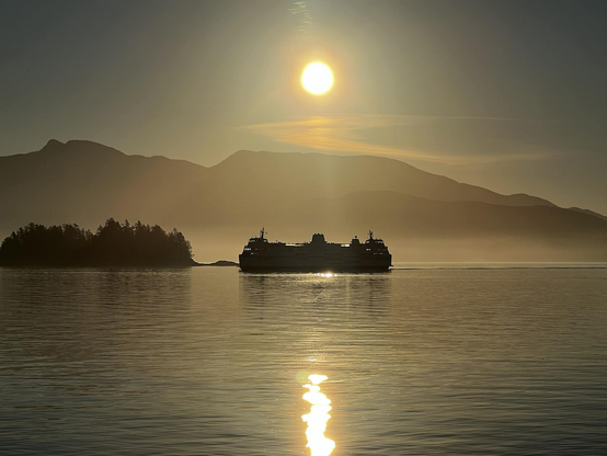 A ferry on calm waters at sunrise with sun reflecting on the water and silhouetted mountains in the background.