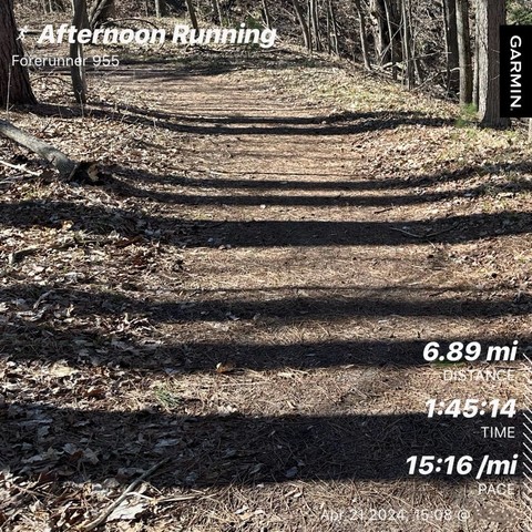 A trail through a wooded area with running statistics overlay: 6.89 mi distance, 1:45:14 time, 15:16/mi pace. Text reads "Afternoon Running",  "Forerunner 955”, and Garmin. 