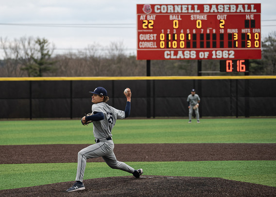 Pitcher wearing a grey shirt with blue highlights holds ball behind himself with outfielder in background as well as a scoreboard thatn Indicates Yale has made 3 runs so far against Cornell's 1.