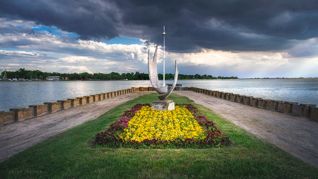 This photo was taken during my visit to Subotica and Palic lake, which is located in the north of the Serbian province Vojvodina. It was one late afternoon in spring. That may day was beautiful, until suddenly storm clouds began to gather over the lake but There was still a little sunlight under the clouds. I was walking along the promenade by the lake when I arrived at this place. I immediately liked what I saw and of course, I took the opportunity to take a shot.

This is a view of Palic lake under a cloudy sky from a small peninsula called "Cape of Poetic Hope". In the distance, you can see the other lake shore and the part of the Palic nature park with its contents. In the central part, surrounded by grass and flowers there is the sculpture "Wings". which is a work of the first academic sculptor from Subotica, Ana Beslic, whose works adorn Palic and Subotica. This sculpture from 1957. was moved from the site of the Memorial Fountain in 1981. to its current position.
The sculpture "Wings" is certainly one of the sights of Palic lake and you just can't miss it if you walk by the lake shore.
Palic lake with its contents and sights is one of the main touristic attractions in this region and you should surely visit it if you ever come to Serbia.

One May day. Palic, Vojvodina, Serbia. From the Monuments and Sculptures collection.
