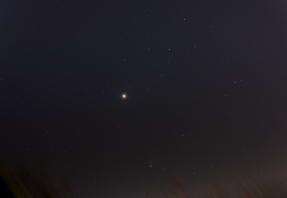 A noisy, heavily processed night sky scene, with blurry artifacts around the horizon from foreground object. In the center just above the horizon is the comet, which appears as a relatively bright smudge with a clearly visible tail pointing almost straight up. Jupiter is a bright disk near the center of the image, slightly to the left of the comet, surrounded by a ragged halo