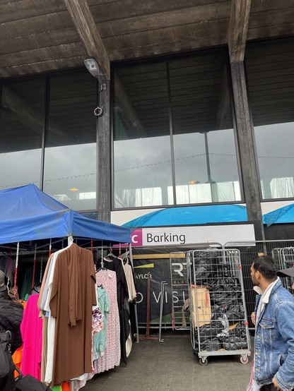 Sign for Barking station on the outside of the building. There are market stalls and people bustling by
