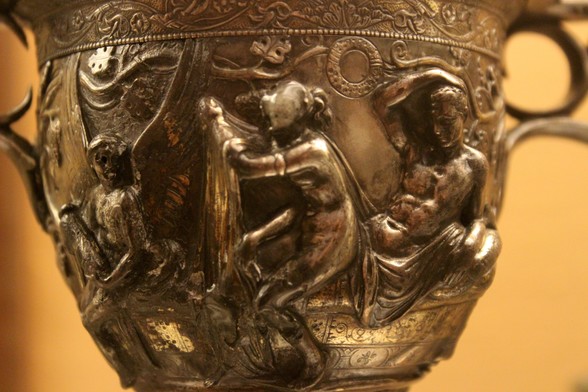 Relief on a silver kantharos cup depicting the love of Venus and Mars. Both are naked, he is reclining on a couch while she sits, facing away from him, on his thighs, getting undressed with the fabric of her dress in hand.
A winged Eros holding a burning torch symbolises their passion.