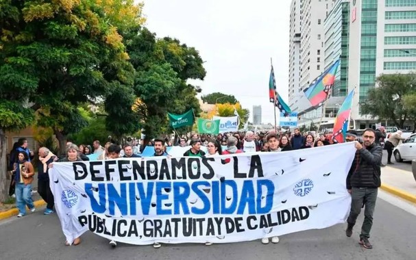 Foto que muestra una marcha. Una multitud de gente caminando por la calle de una ciudad, con árboles y edificios de fondo, y al frente se observa una bandera que lleva la gente y dice: Defendamos la Universidad pública, gratuita y de calidad.