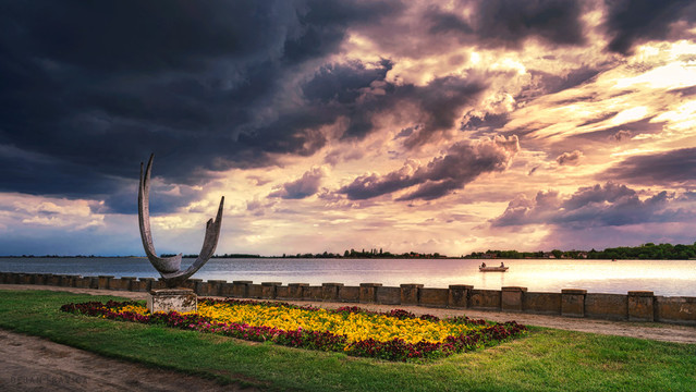 I took this photo one beautiful may day during my visit to Subotica and Palic lake, which is located in the north of the Serbian province Vojvodina. That spring day was beautiful until suddenly storm clouds began to gather over the lake but There was still a little sunlight under the clouds. I was walking along the promenade by the lake when I arrived at this place. I liked this scene so I took a shot and moved on.

This is a view of Palic lake under the dramatic cloudy sky from a small peninsula called "Cape of Poetic Hope". In the distance, you can see the other lake shore and the part of the Palic nature park and on the left, surrounded by grass and flowers there is the sculpture "Wings". which is actually located in the central part of this small peninsula. On the lake, not so far from the shore, there is a small boat illuminated with the last sun rays that day.
This sculpture is a work of the first academic sculptor from Subotica, Ana Beslic, whose works adorn Palic and Subotica. In 1957. it was moved from the site of the Memorial Fountain in 1981. to its current position.
The sculpture "Wings" is certainly one of the sights of Palic lake and you just can't miss it if you walk by the lake shore.
Palic lake with its contents and sights is one of the main touristic attractions in this region and you should surely visit it if you ever come to Serbia.

One May day. Palic, Vojvodina, Serbia. From the Monuments and Sculptures collection.