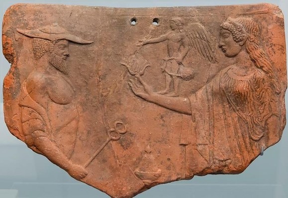 Clay tablet with a relief of Hermes and Aphrodite with a tiny Eros. Hermes is depicted as a mature, bearded man with his traveller's hat and his kerykeion staff in hand. Aphrodite, dressed in flowing robes, holds a flower. On her arm stands a winged Eros with a lyre in his left hand.