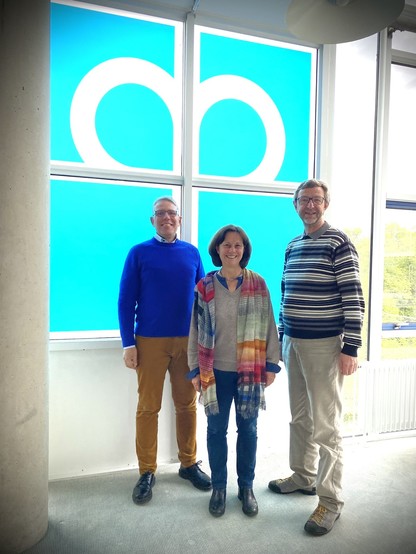 Drei Personen haben sich für Gruppenfoto aufgestellt und schauen freundlich in die Kamera. Im Hintergrund großes Fenster mit Diakonie-Logo: Weißes Kreis auf blauem, quadratischem Grund. In den oberen Feldern des Kreuzes jeweils eine kreisförmige Schlinge