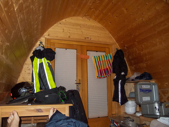 Foto: Blick in einen kleinen Raum mit gebogener Decke, in dem Motorradbekleidung, -koffer und andere Gegenstände angeordnet sind.