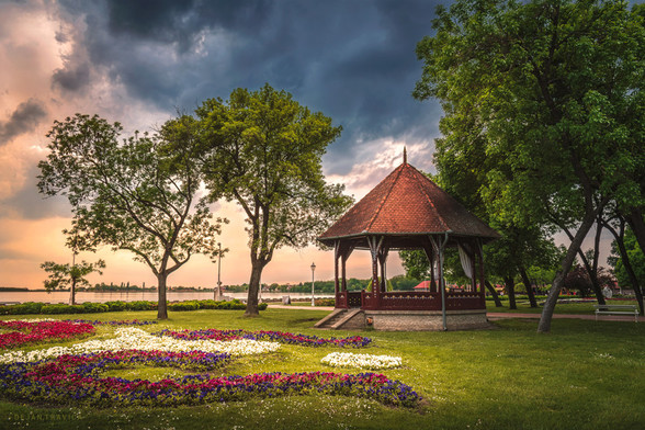 While I was walking along the promenade of the Palic Lake near Subotica in Serbia, one beautiful day in spring I came across this interesting old building which I immediately liked at first sight. It was surrounded by trees and flowers on the green grass, and the beautiful Palic Lake could be seen close to it. The day was coming to end and the sunset was slowly approaching. A storm clouds began to gather in the sky so I had to photograph it quickly and moved on to avoid the rain.

One May day on the Palic Lake, Vojvodina, Serbia. From the Parks and Gardens collection.