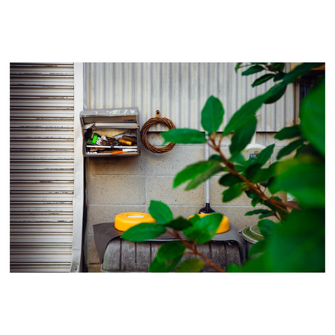This image captures a curious urban scene where a battered metal mailbox, repurposed to store an array of tools, creates a quirky focal point. Next to it hangs a circular wicker wreath on the concrete wall. Below, yellow barbell weights and cleaning equipment rest casually, creating a spontaneous composition. A glimpse of green leaves in the foreground introduces a touch of nature to this eclectic tableau set against the industrial backdrop of corrugated metal and concrete.