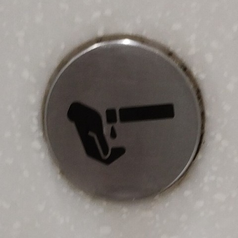 A metal disk mounted on the wall has a black icon on it. The icon has three elements:
• a hand.
• a vertical bar, in 2 parts.
• a droplet of liquid.

The hand is to the left of the bar. With the fingers slightly under the 2-part bar. A droplet from the 2-part bar would logically end up in those fingers.

The two-part bar looks ode tocal to the two-part bar in No Smoking or Smoking icons: a long bar with a tiny square (or ash) at the tip of the cigarette.

In this icon, the bar represents the visible part of the dispenser, which happens to be shaped like a cylindrical bar. It juts out from the wall below the icon (not shown).
