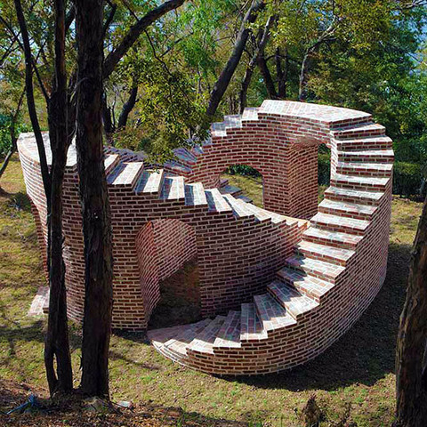 Photograph of a sculptural work of art by Michel de Broin. The image is looking down on what amounts to two brick staircases intertwined such that as you walk down one curving stairway, you exit under the other staircase and the path goes right back up the other side. Reminds me a little of an Escher-style drawing. Very practical, and yet quite whimsical, the brick staircases sit in a grassy area amongst some trees in what I imaging must be a larger garden of other sculptural works—perhaps a park-like setting?