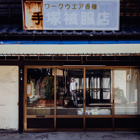 The image presents a quaint storefront with a large, faded sign above it that has suffered from significant paint erosion, lending it a rustic charm. Through the glass front of the store, we can see various items on display, suggesting the shop's commercial history. A photographer is subtly reflected in the glass, adding a layer of narrative about the act of capturing images. The shop and its sign are symbols of the city's working past, standing against the test of time.