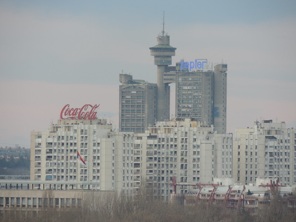 a photo of the building from far away, it is towering over other buildings, there is a large red coca-cola sign on a white building in front of it, it is cloudy
