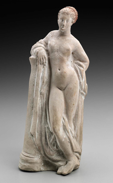 Statuette of Aphrodite leaning on a column. There are traces of red paint in her elaborately braided hair. She is naked, only a string of pearls or beads gathering in her cleavage and decorating her breasts.