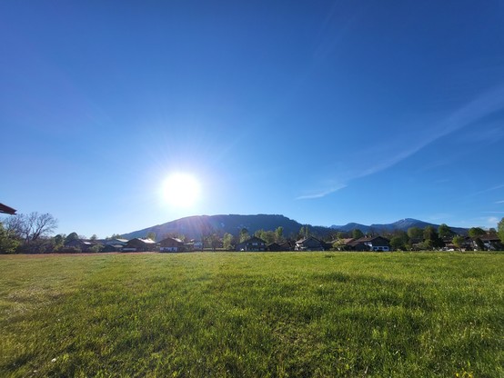 A serene and picturesque scene captured in this image, showcasing a vast green field with houses dotted in the distance and majestic mountains towering in the background. The sky above is a clear blue, adding to the overall tranquility of the landscape. The sunlight gently bathes the field, creating a warm and inviting atmosphere. The grass sways gently in the breeze, hinting at a peaceful morning setting. This rural area exudes a sense of calm and tranquility, making it a perfect escape from t…
