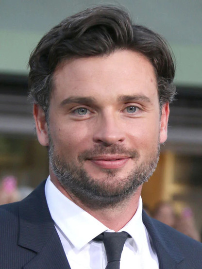 Portrait de Tom Welling. Il porte un costume gris foncé avec une chemise blanche et une cravate gris foncée. Il regarde directement l’objectif.