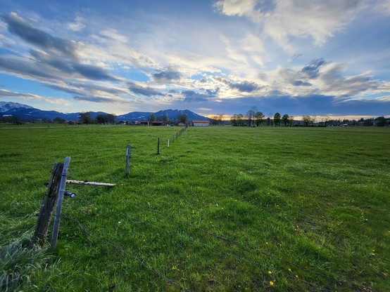 A serene scene of a lush green field under a clear blue sky, with fluffy white clouds floating overhead. In the foreground, a sturdy wooden fence stretches across the landscape, dividing the field into sections. The grass is vibrant and abundant, swaying gently in the breeze. The image exudes a sense of tranquility and natural beauty, with the fence adding a touch of rustic charm to the rural setting. This idyllic countryside landscape is perfect for those seeking a peaceful escape into nature,…