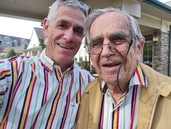Photo: A selfie of two men with graying hair, smiling. Both are wearing similar white shirts with vertical blue, yellow, red and green stripes. One man is also wearing glasses and a light beige jacket. There is a low apartment building in the background. A number of windows are showing in the background.