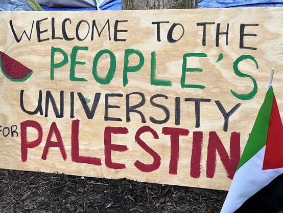 Sign on a board at Harvard solidarity encampment. It says “Welcome to the People’s University for Palestine. “ there is a watermelon painted on the left side of the sign and Palestinian flag planted in the ground in front of the right side of the sign. 