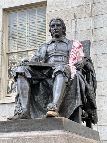 Picture of the statute of John Harvard, a patrician looking young man in a seated position, draped with a red keffiyeh. 