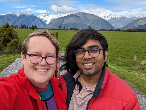 Selfie of a gay couple (Mark and Dee) with mountains, snow, and Fox Glacier in the background. Mark squints at the camera with a force smile, while Dee has an effortless look of contentment and long hair swept to one side that almost seems carried in the breeze. Both are wearing tortoiseshell-style glasses; Mark's are pinkish and Dee's are bluish. Both are wearing red coats. Undearneat Mark has a colourful jumper while Dee has a pinkish collared t-shirt.