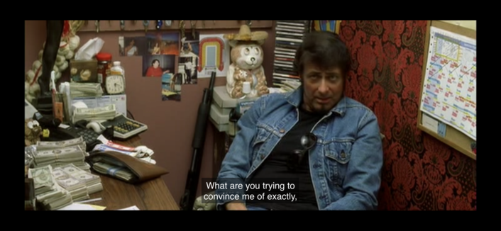 Image from “Kill Bill” showing the image of the owner of the strip club, a masc presenting persons with light skin and dark hair sitting behind desk and wearing a denim jacket
Subtitled: “What are you trying to convince me of exactly”