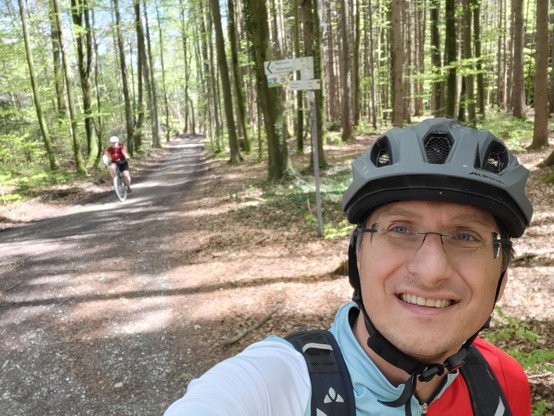 A man in a forest is taking a selfie with a bicycle helmet on his head. The man is wearing glasses and appears to be smiling. The dominant colors in the image are grey, with an accent color of red. The man, who is in his mid-40s, is the main focus of the image, with the background consisting of trees and foliage. The man is standing on the ground, surrounded by nature. He is wearing casual clothing and appears to be enjoying his time outdoors. The image captures a moment of outdoor recreation, …