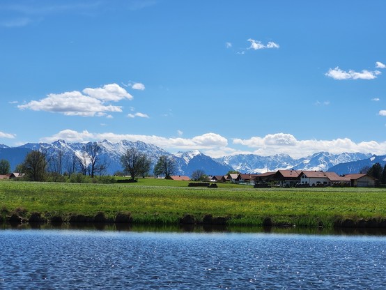 A picturesque scene of a tranquil lake surrounded by lush green grass and trees, with charming houses dotting the landscape. In the background, majestic mountains rise against a clear blue sky with fluffy white clouds. The water of the lake reflects the beauty of the surroundings, creating a serene and peaceful atmosphere. Two trees stand tall in the foreground, adding to the natural beauty of the scene. This idyllic outdoor waterside setting is perfect for relaxation and enjoying the beauty of…