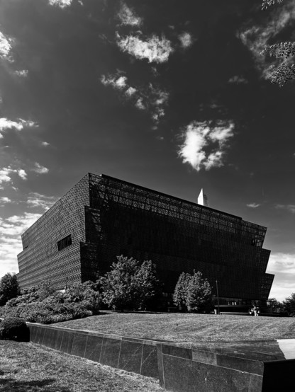 Black and white wide picture of the African American Museum with a piece of the Washington monument poking out on the Mall in DC