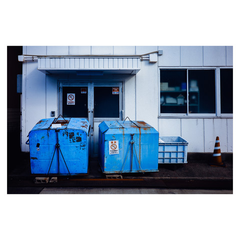 This vibrant photo features a stark white building with a row of vivid blue bins lined up against it. The bins, bearing signs of wear and use, stand out against the cleanliness of the building's facade, hinting at the function and form of city life. A solitary traffic cone adds a pop of orange to the scene, complementing the blues and whites.