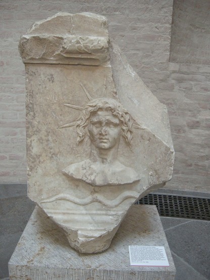 Relief of a bust of Helios on a white marble altar. The bust is placed above a snake. Helios is depicted with chin-length curly hair and a seven-spiked sunray crown, part of which has broken off on the right side.